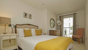 A bedroom with a bed, bedside table, lamp, mirror, chair, and a window with curtains. - Stylish comfortable apartment in central Kingston - Pass the Keys (Kingston-upon-Thames)