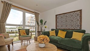 A living room with a green sofa, a round coffee table, and a chair. There is a window with curtains, a decorative wall panel, and a view of buildings outside. - Stylish comfortable apartment in central Kingston - Pass the Keys (Kingston-upon-Thames)