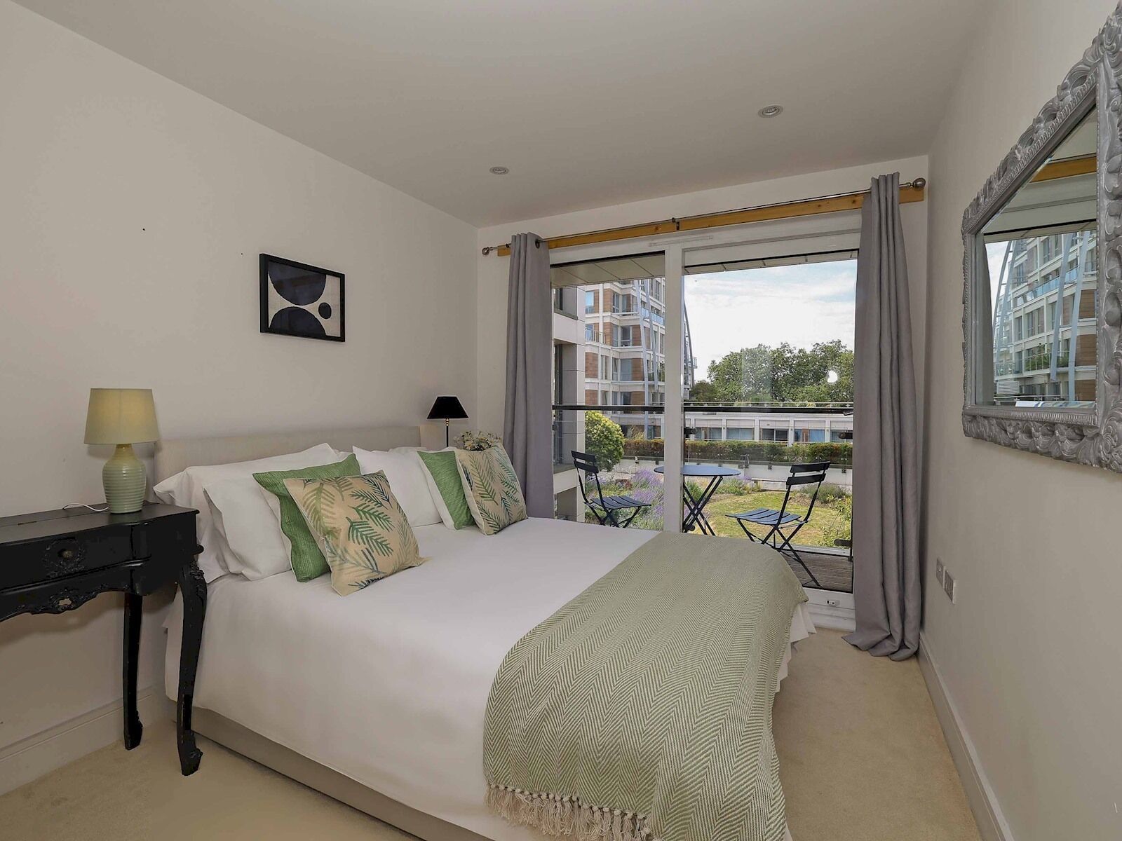 A bedroom with a bed, a desk, a chair, a window with curtains, and a view of buildings and greenery.
