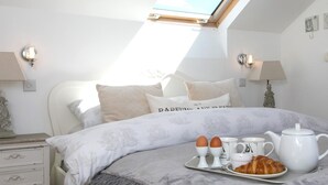 A bedroom with a skylight, a bed with a floral bedspread, bedside lamps, a dresser, and a breakfast tray with eggs, coffee cups, and a croissant. - Beautiful 3 bedroom apartment with Pool Access - Pass the Keys (Bae Trearddur)