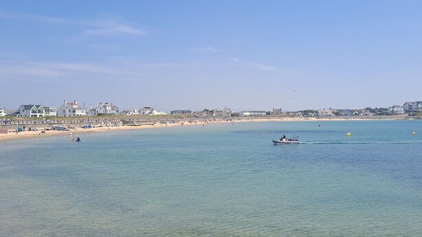 A coastal beach scene with clear water, a sandy shore, and a row of houses in the background. - Beautiful 3 bedroom apartment with Pool Access - Pass the Keys (Bae Trearddur)
