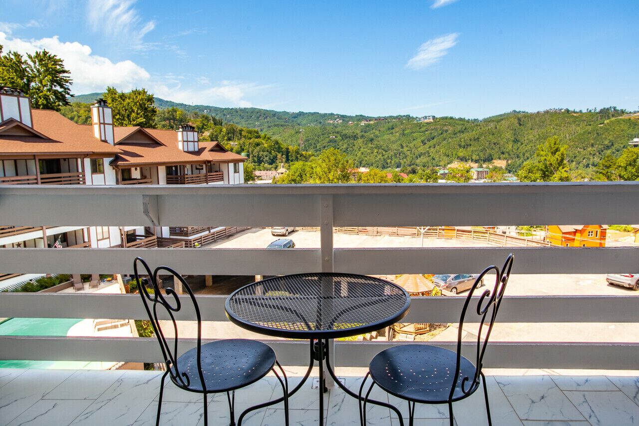 A balcony with a round table and two chairs, overlooking a residential area with houses and a hilly landscape.