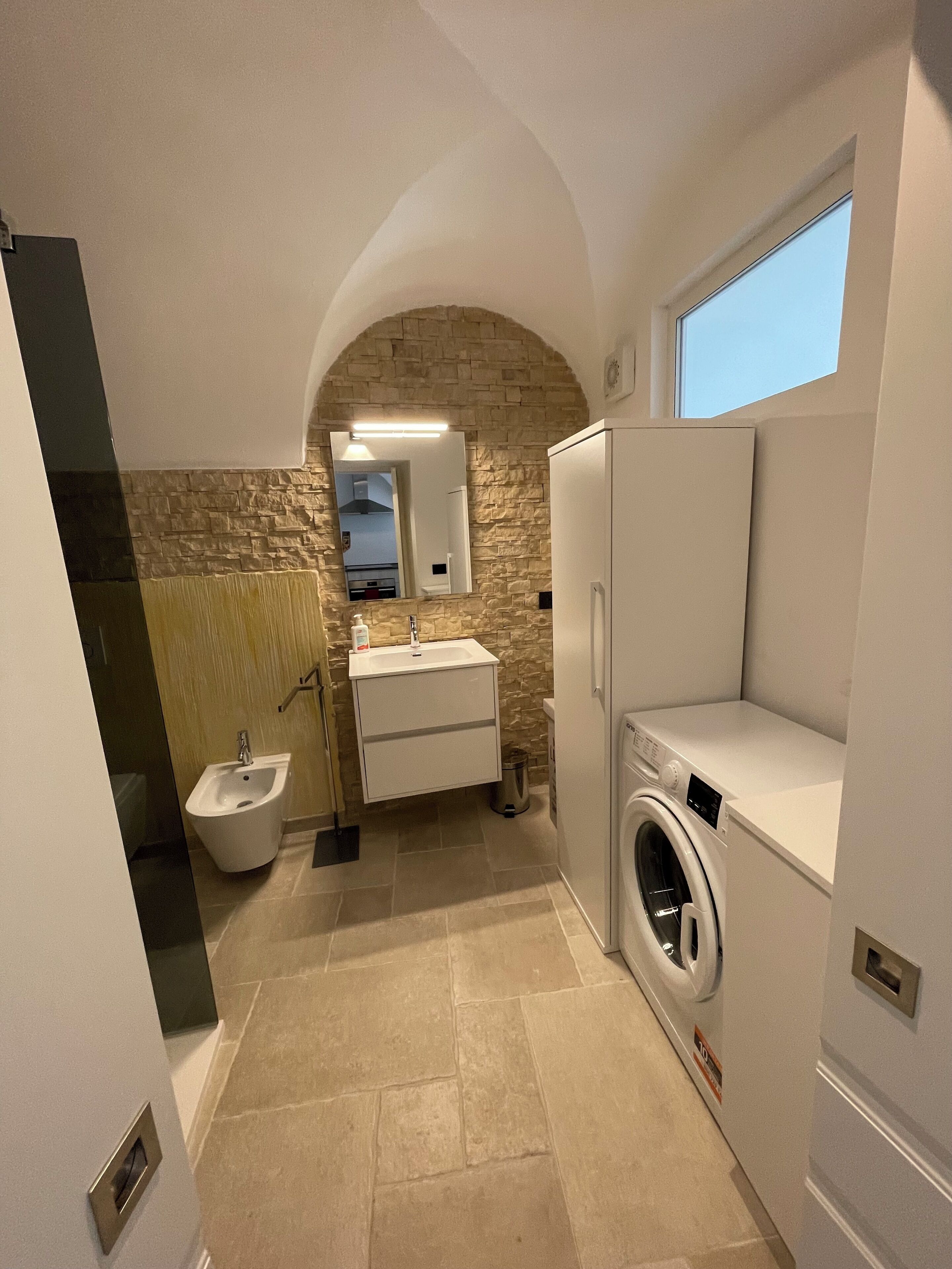 Une salle de bain compacte avec un mur d’accent en pierre, un lavabo, un bidet et un lave-linge.