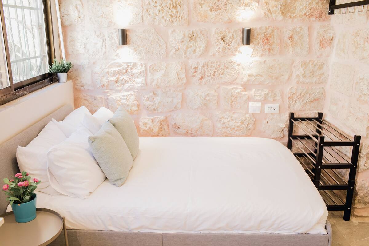 Un dormitorio con pared de piedra, cama con ropa de cama blanca, una mesita con planta en maceta y una silla.
