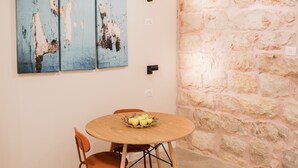 Un comedor con una mesa redonda de madera, dos sillas naranjas y un frutero. Hay tres cuadros azules en la pared y una pared de piedra a la derecha.