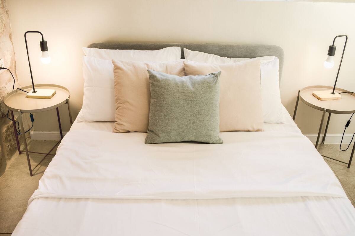 Una cama bien hecha con ropa de cama blanca y gris, dos mesitas de noche con lámparas y una pared lisa.