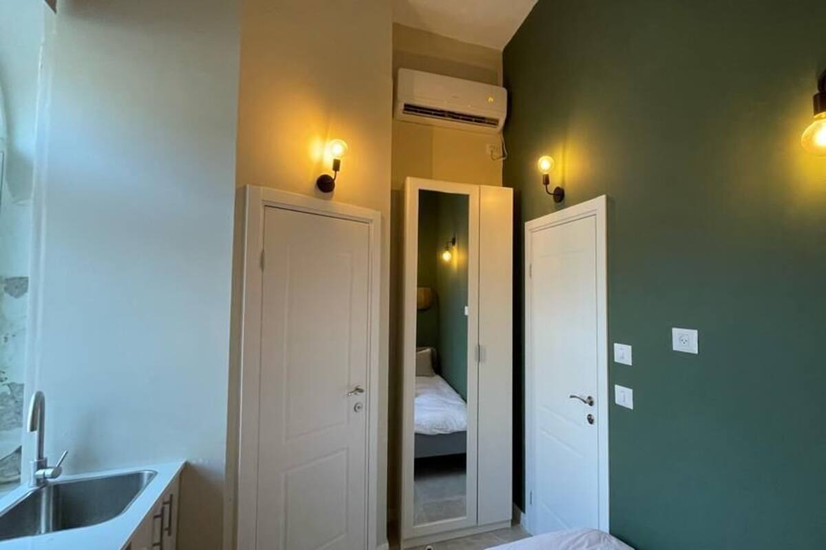 Una habitación con una cama, una puerta blanca, un lavamanos y una lámpara fijada a la pared.