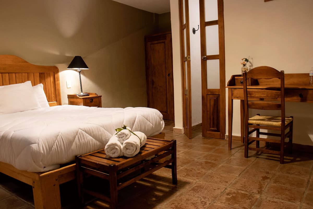 A neatly made bed with white linens, a wooden nightstand with a lamp, and a bench with rolled towels.