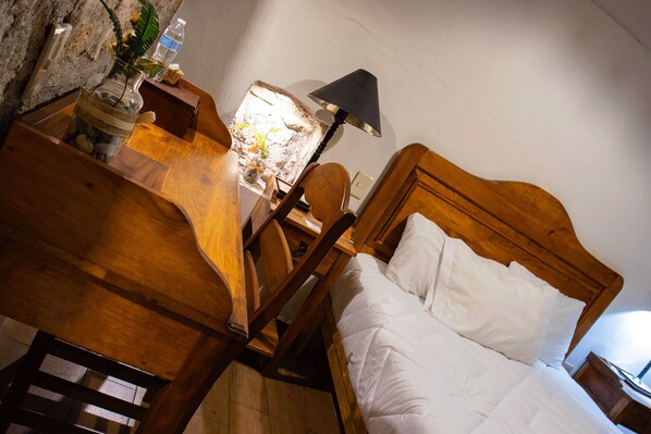 Una cama de madera con ropa de cama blanca, una mesita de noche de madera con una lámpara, y una pared de piedra.