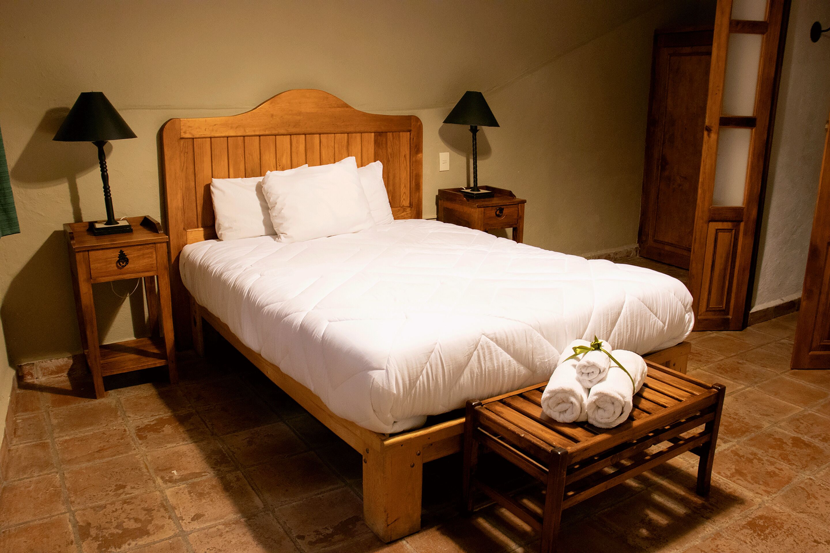 Un lit bien fait, avec du linge de lit blanc, deux tables de chevet avec des lampes et un banc en bois sur lequel sont posées des serviettes.