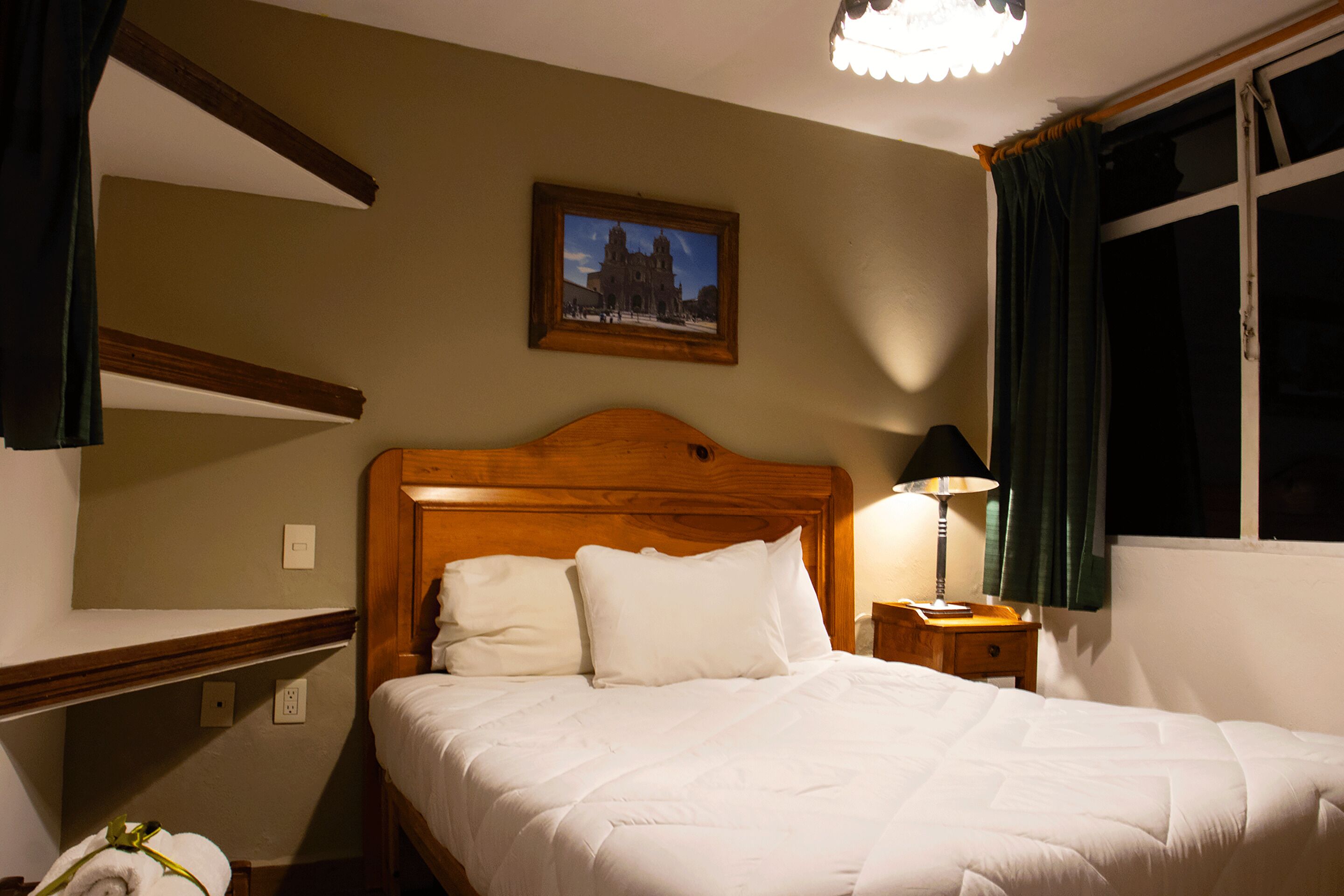 A neatly made bed with white linens, a wooden headboard, a nightstand with a lamp, a framed picture on the wall, and a window with curtains.