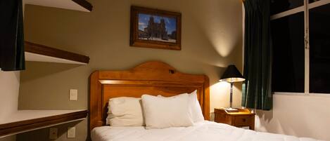 A neatly made bed with white linens, a wooden headboard, a nightstand with a lamp, a framed picture on the wall, and a window with curtains.