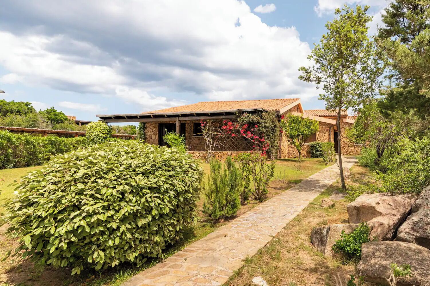A stone house with a tiled roof, surrounded by a well-maintained garden with various shrubs and a stone pathway leading to the entrance.