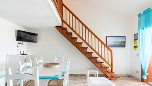 A dining area with a wooden staircase, white chairs, a table with a bowl, and a television mounted on the wall. - Cala Paradiso Apt "Casa Agave" ⭐⭐⭐⭐ Sea View, Garden, WiFi, AC (Salina Bamba)