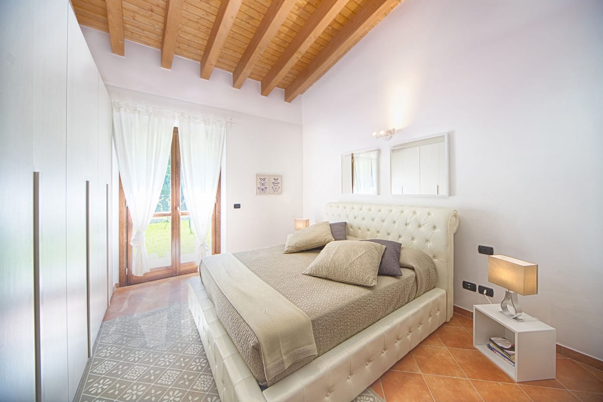 A bedroom with a large bed, a nightstand, a window with curtains, and a view of the outdoors.