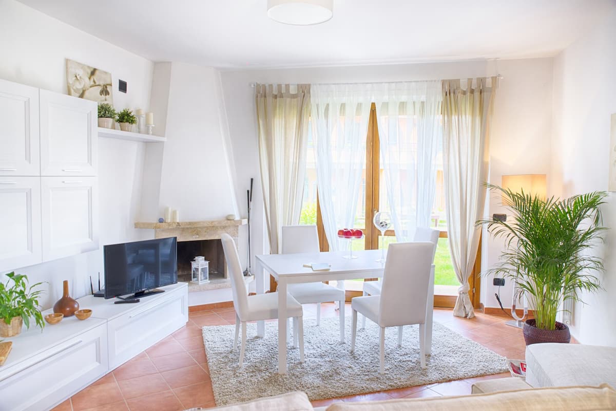 A modern living room with a TV, white furniture, a fireplace, and a large window with curtains.