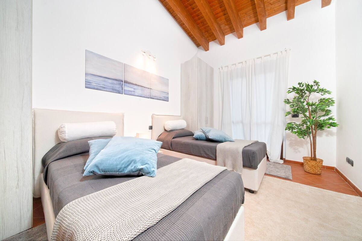 A hotel room with two beds, a large window with curtains, a ceiling with exposed beams, and a potted plant.
