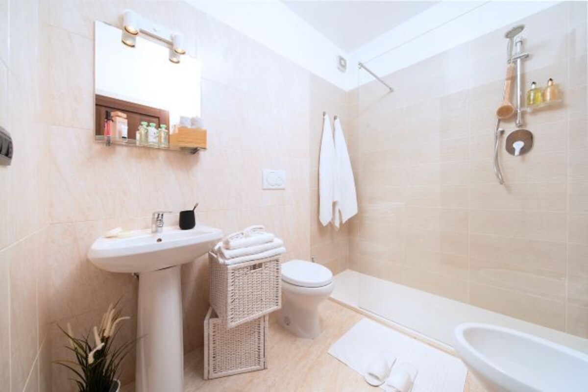 A modern bathroom with a pedestal sink, toilet, and bathtub. There is a mirror with lights above it, and a shelf with toiletries.