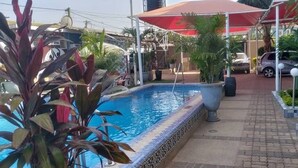 A swimming pool with a tiled edge, surrounded by potted plants and a covered walkway. - Sweet Mama Residence  (Serrekunda)