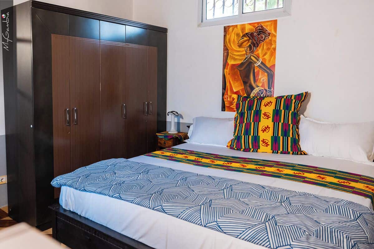 A bedroom with a large wardrobe, a bed with patterned bedding, a colorful pillow, and a painting on the wall.