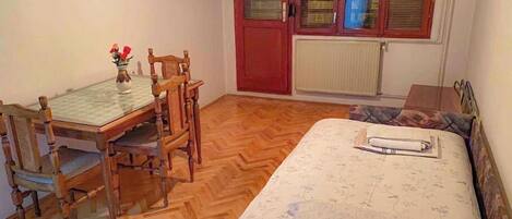 A small, cozy room with a wooden herringbone floor, a single bed with a floral bedspread, a wooden dining table with two chairs, and a small table with a vase of flowers.