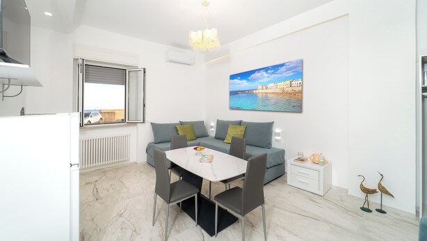 Ein modernes Wohnzimmer mit Esstisch, grauen Sofas, einer weißen Kommode und einem Gemälde, das eine Küstenlandschaft zeigt.