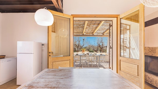 A compact kitchen with a white refrigerator, a wooden dining table, and a view of the outdoors through an open door. - 3621 Tenuta Terra del Sole by Perle di Puglia (Casarano)
