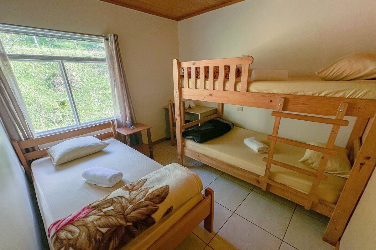 Une chambre avec un lit simple, un lit superposé, une fenêtre avec des rideaux, une petite table et une chaise.