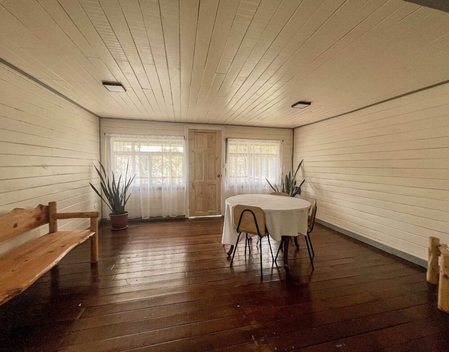A room with a wooden floor, a round table with chairs, a bench, and two potted plants.