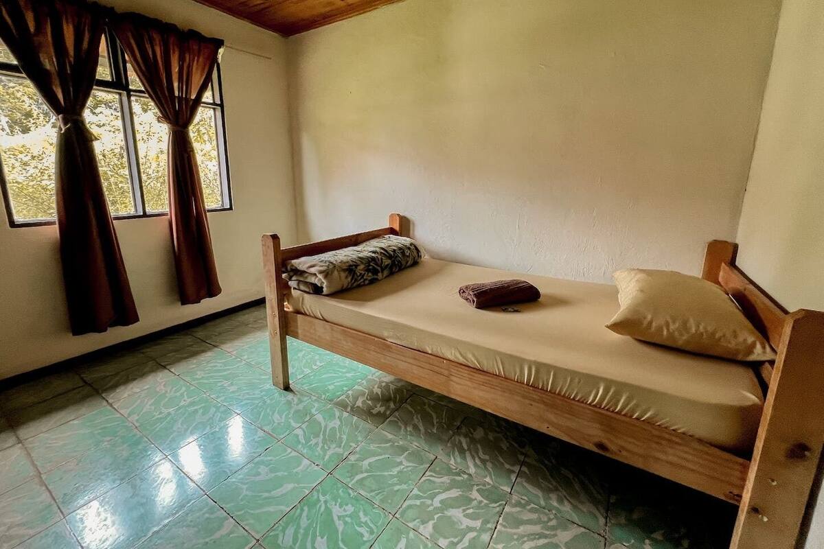 Une petite chambre avec un lit simple, un cadre en bois, un matelas et un oreiller. Il y a une fenêtre avec des rideaux, et le sol est carrelé.