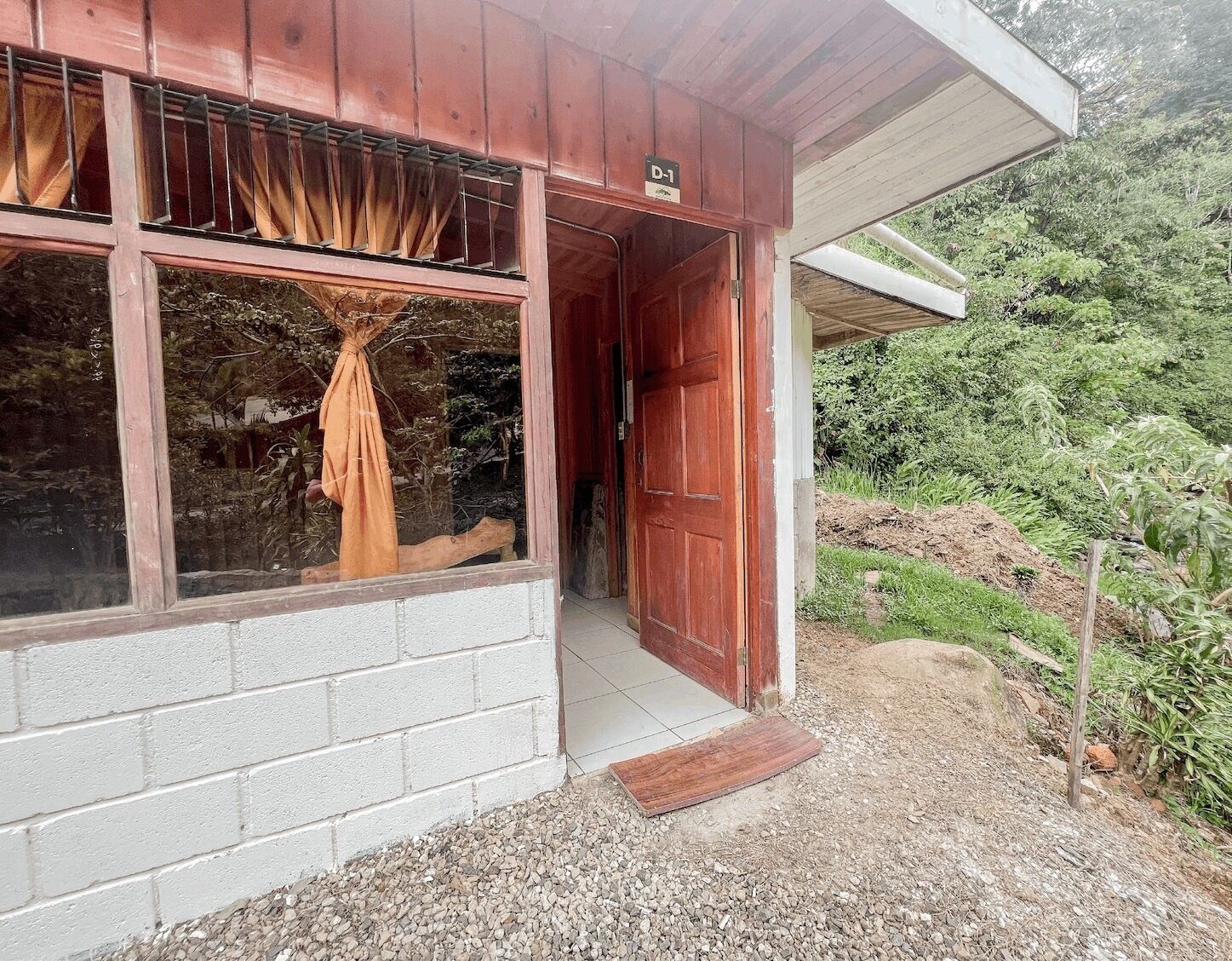 Un edificio rústico de madera con un ventanal grande y una puerta, rodeado de follaje.