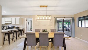 A modern kitchen with white cabinets, a dining area with a wooden table and grey chairs, and a view of a patio through glass doors. - The EBB TIDE-private pool + just mins to beach! (Palm Coast)