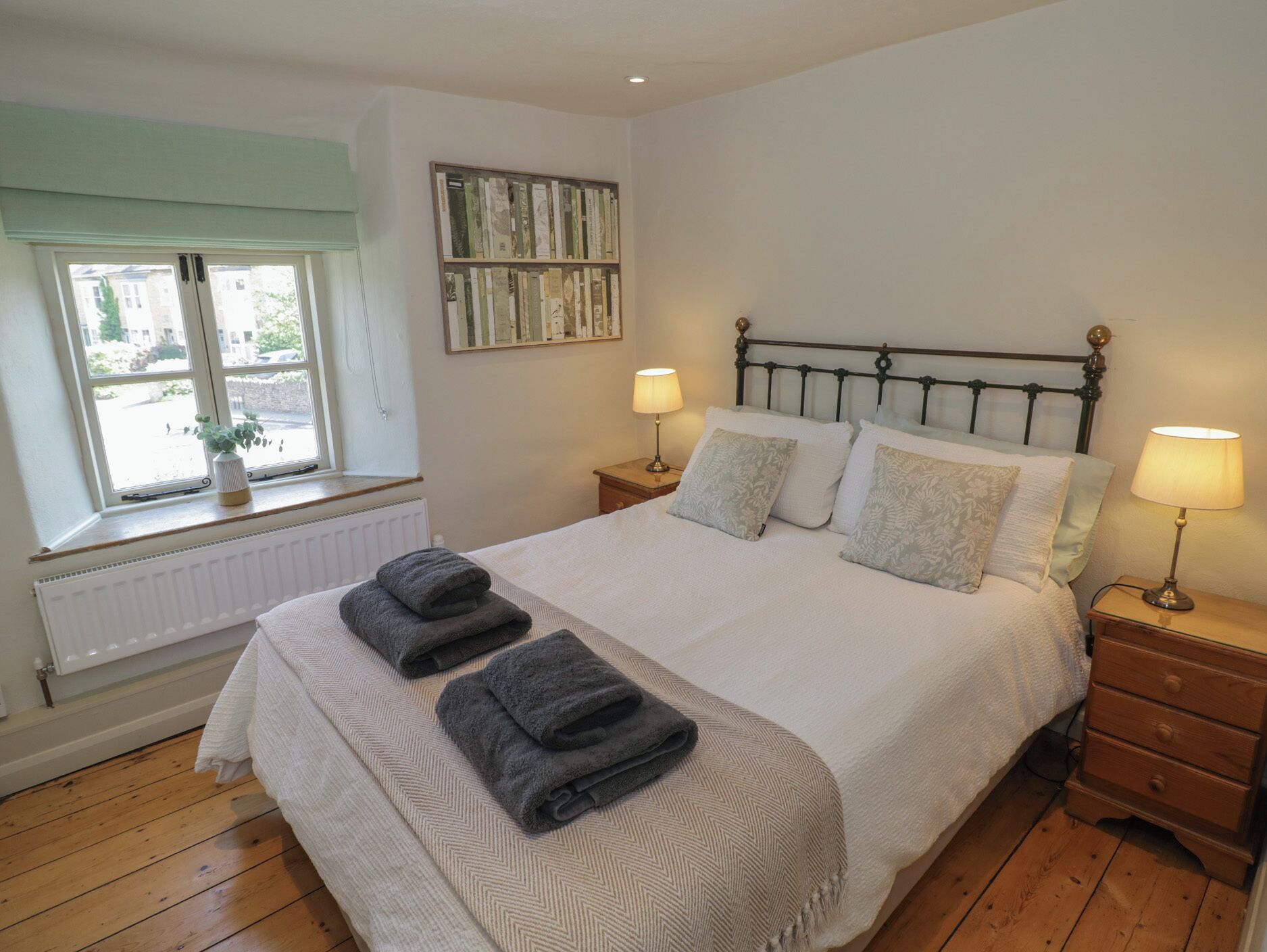 A neatly made bed with two folded towels, a bedside lamp, a nightstand, a window with a view, and a bookshelf.