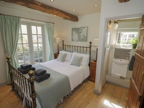 A bedroom with a bed, a window with curtains, a bedside table with a lamp, a wooden floor, and a view of the outside.