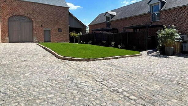 A courtyard with cobblestone flooring, a green lawn, and brick buildings with a garage and covered patio area. - Bienvenue à la Ferme (Maresches)