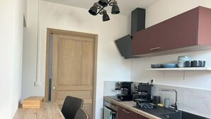 A compact kitchen with a wooden dining table, black chairs, a built-in oven, and a coffee machine. - Bienvenue à la Ferme (Maresches)