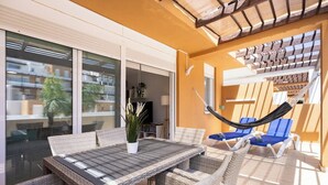 Un espacio al aire libre iluminado por el sol con una mesa de madera, sillas de mimbre, una hamaca y sillones azules.