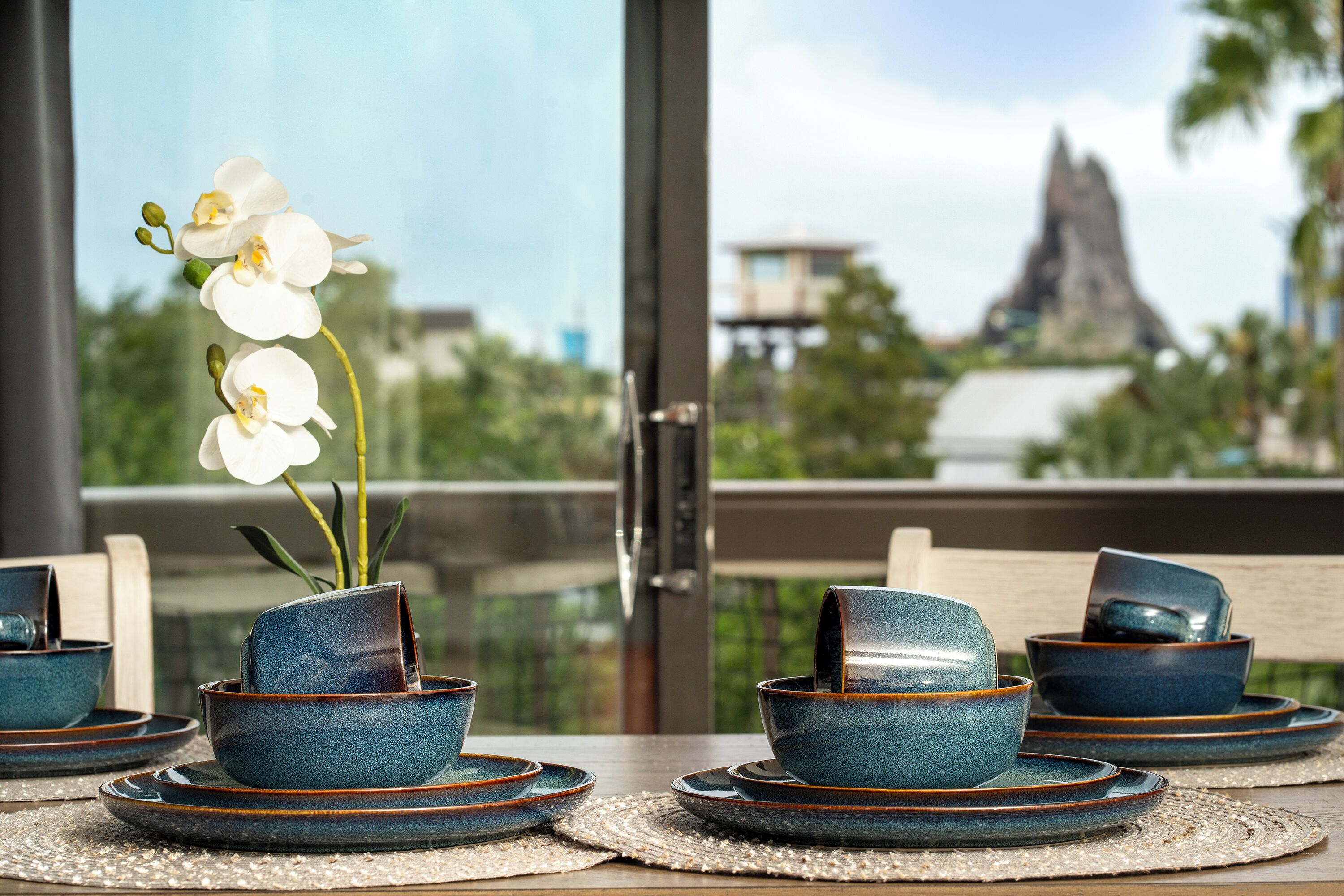 A dining table with blue ceramic bowls and plates, a vase with white flowers, and a view of a mountain through a glass door.