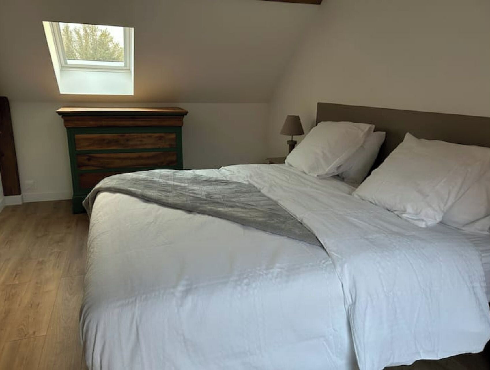 Une chambre à coucher comprenant un lit, une table de chevet, une lampe, une commode et une lucarne.