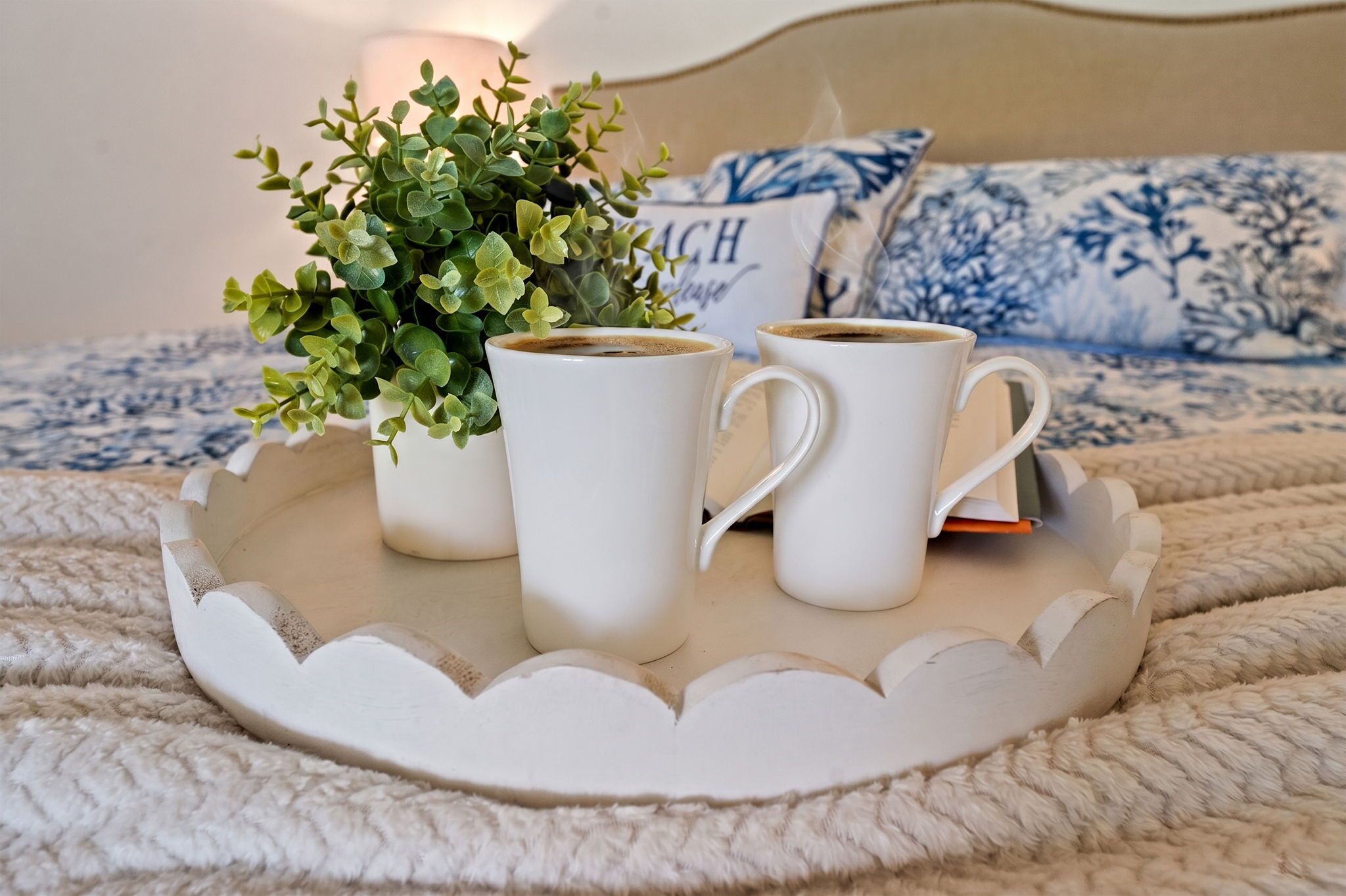 Un plateau avec deux tasses à café blanches, une petite plante verte et un coussin à motifs bleus sur un lit.
