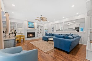 A spacious living room with a fireplace, a blue sofa, and a kitchen area in the background.