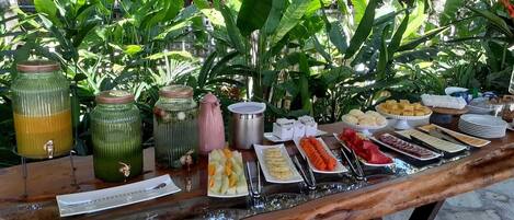 Una mesa de buffet con diversos alimentos y bebidas, rodeada de abundante vegetación.