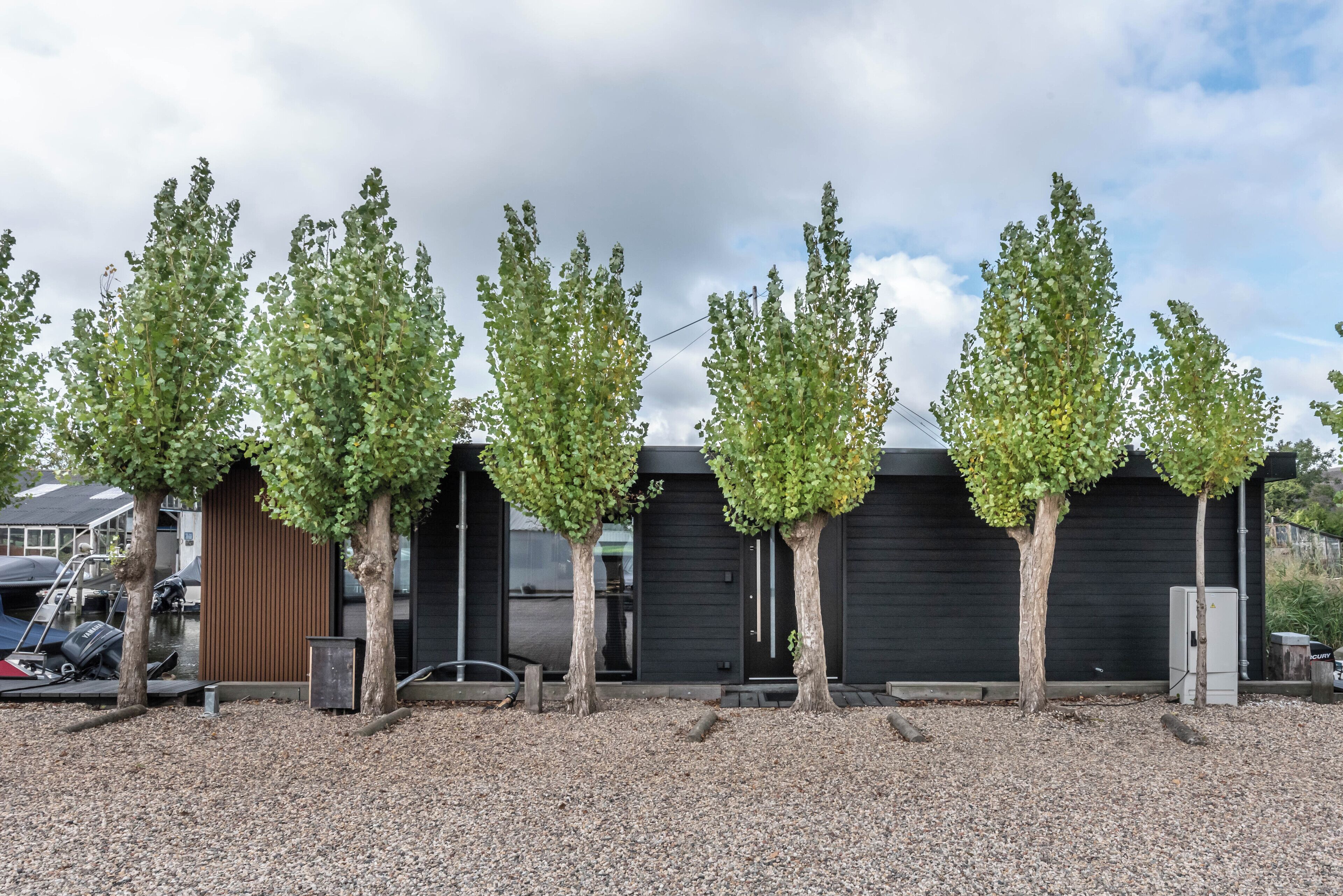 Une rangée d’arbres se trouve devant un bâtiment à la façade noire et à l’allée gravillonnée.