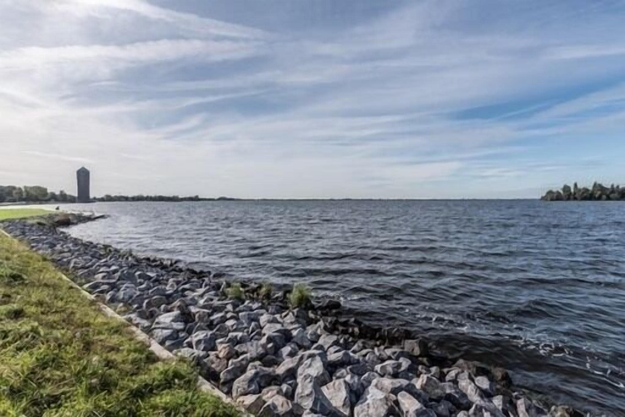 A waterfront view with a rocky shoreline, a grassy area, and a distant building.