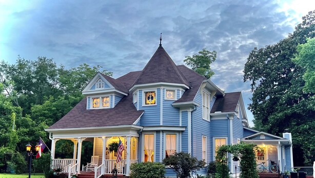A large, two-story house with a wrap-around porch, a gabled roof, and a small tower-like structure on the roof.