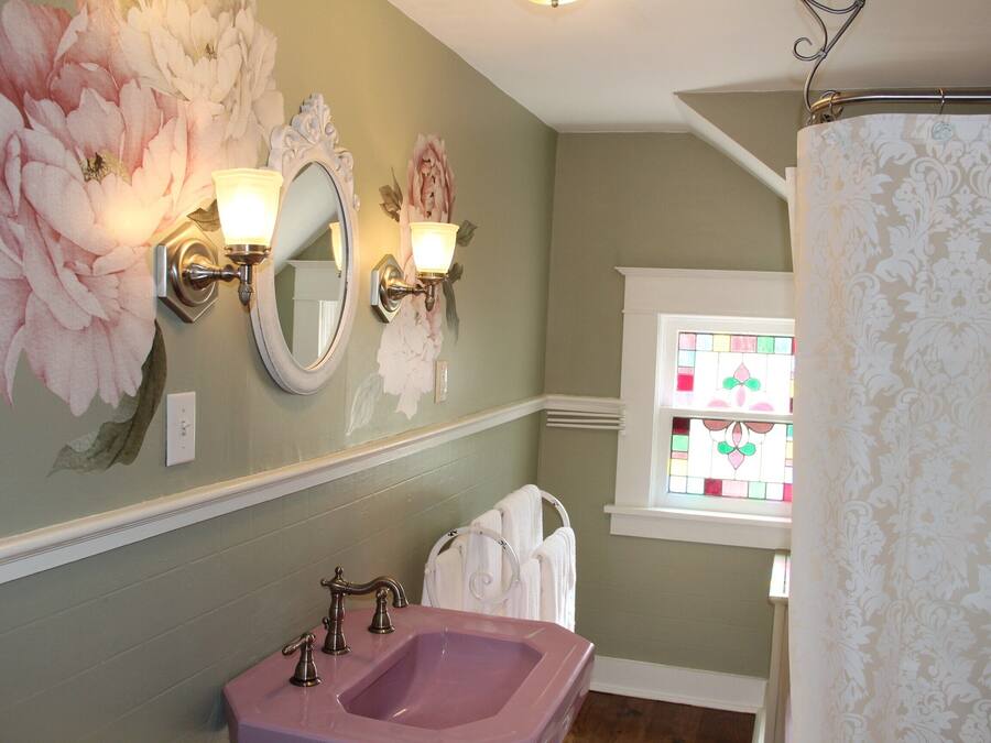 Une salle de bain avec un lavabo rose, un miroir agrémenté d’appliques murales et une fenêtre avec des vitraux.