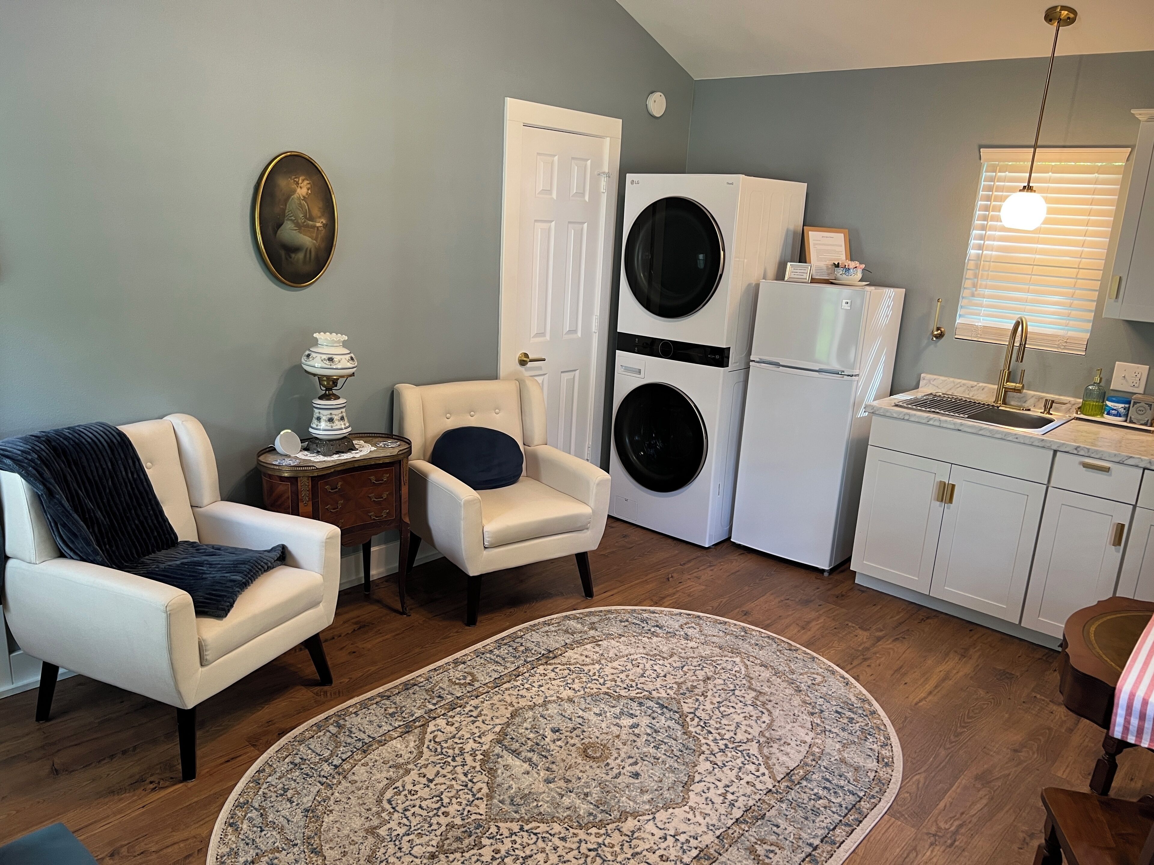 A compact living space with a beige armchair, a round rug, a small side table with a lamp, a washing machine, a refrigerator, and a kitchen area with a sink and window blinds.