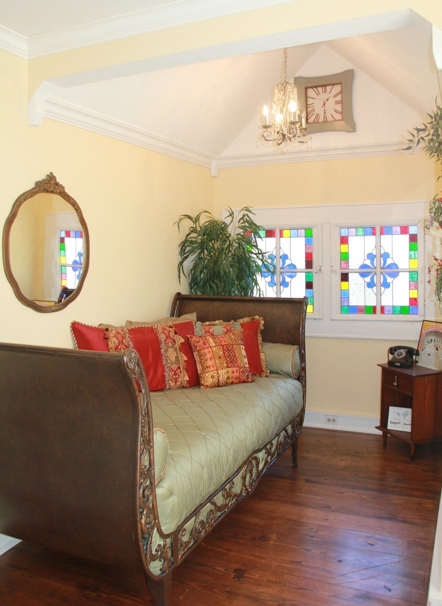 A cozy room with a large bed, a mirror, a chandelier, and a window with stained glass.
