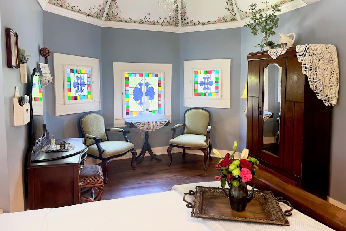 A room with a curved ceiling, three chairs, a small table, a wooden cabinet, and a bed with a white bedspread.