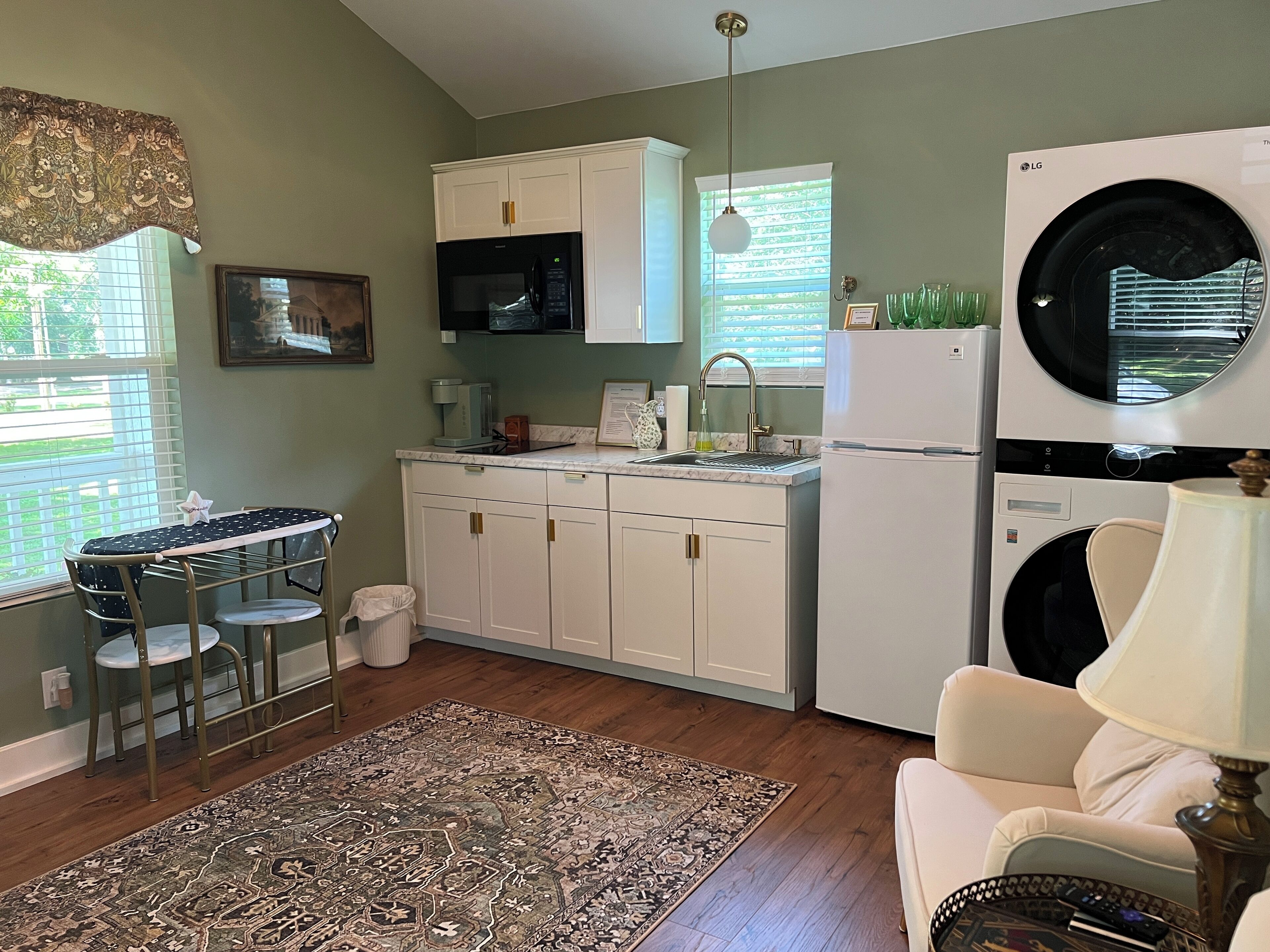 A compact kitchen with white cabinets, a microwave, a sink, and a washing machine. There is a dining table with chairs, a patterned rug, and a sofa with a lamp.